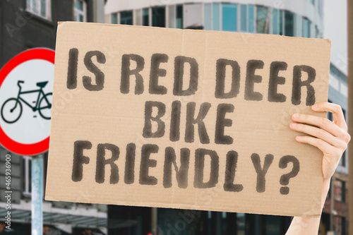 The question " Is Red Deer bike friendly? " on a banner in men's hand with blurred background. Transportation. Zero waste. Bicycle lane. Streets. City. Safety. Insecure. Road signs. Dangerous