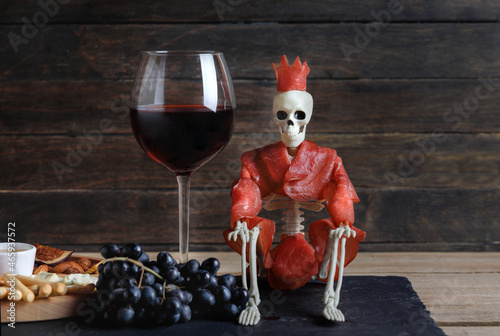 Halloween Skeleton In Bresaola Meat Sitting With Glass Of Red Wine In Glass On Wooden Background. Side View.