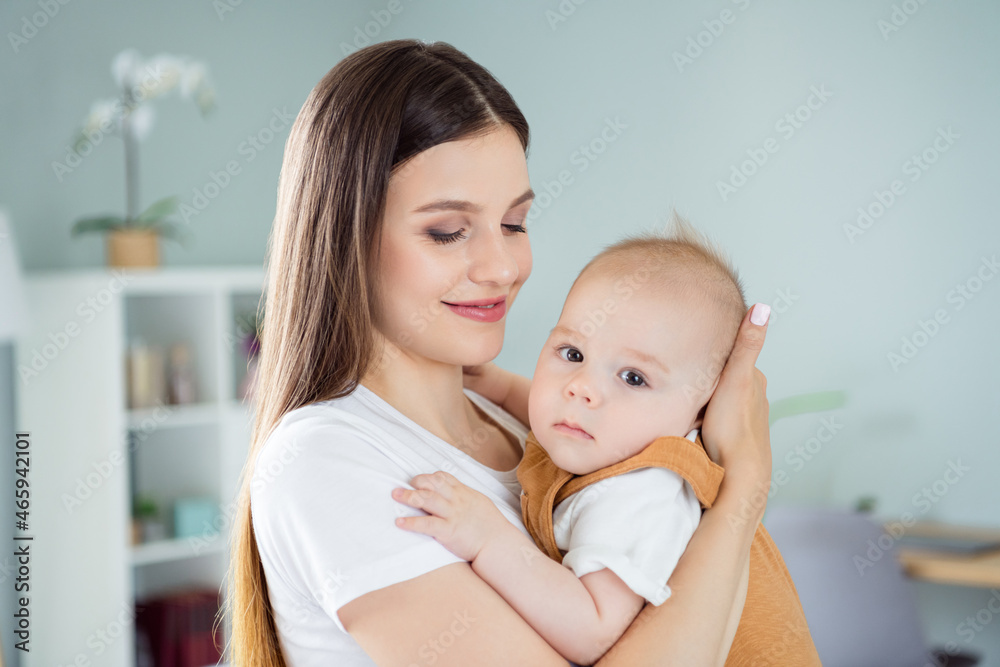 Photo of happy joyful dream family mom cuddle little small infant kid look camera wear white t-shirt home indoors Photo of happy joyful dream family mom cuddle little small infant kid look camera wear white t-shirt home indoors