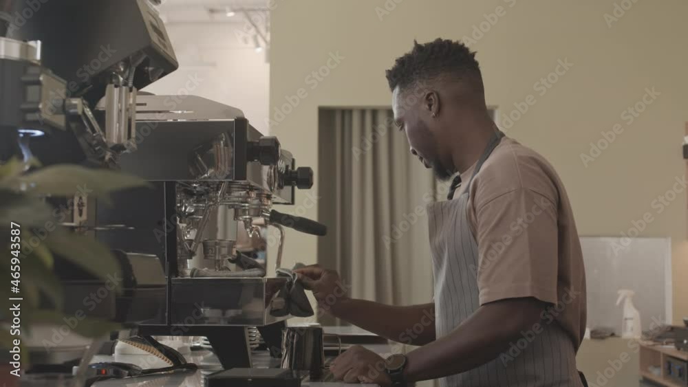Side view of young Black male barista wearing apron, cleaning coffee machine in morning, wiping it with rag Side view of young Black male barista wearing apron, cleaning coffee machine in morning, wiping it with rag