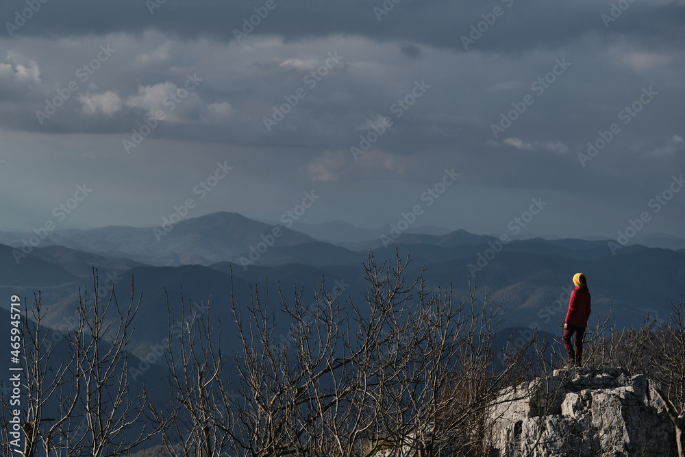 View from back of tourist in nature in mountains. Traveler girl in red jacket and bright yellow hat stands on top of cliff and enjoys views of mountains in distance.