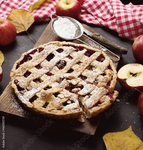 baked round traditional apple pie on brown wooden board and fresh red apples, top view