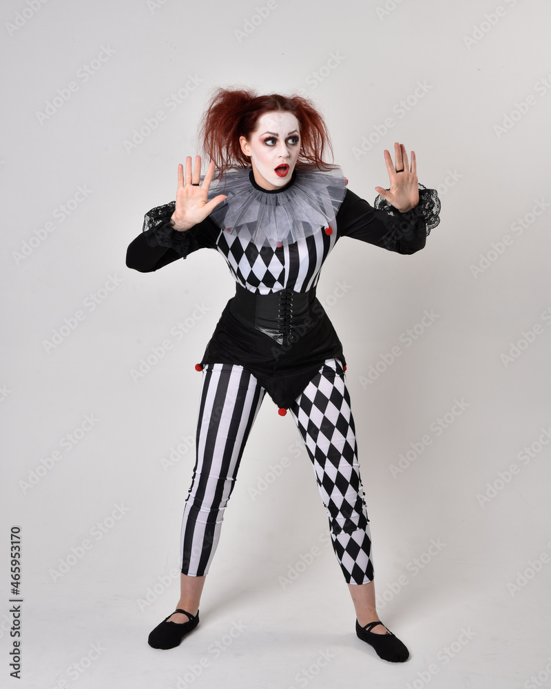 Full length portrait of red haired girl wearing a black and white clown jester costume, theatrical circus character. Standing pose isolated on studio background. Full length portrait of red haired girl wearing a black and white clown jester costume, theatrical circus character. Standing pose isolated on studio background.