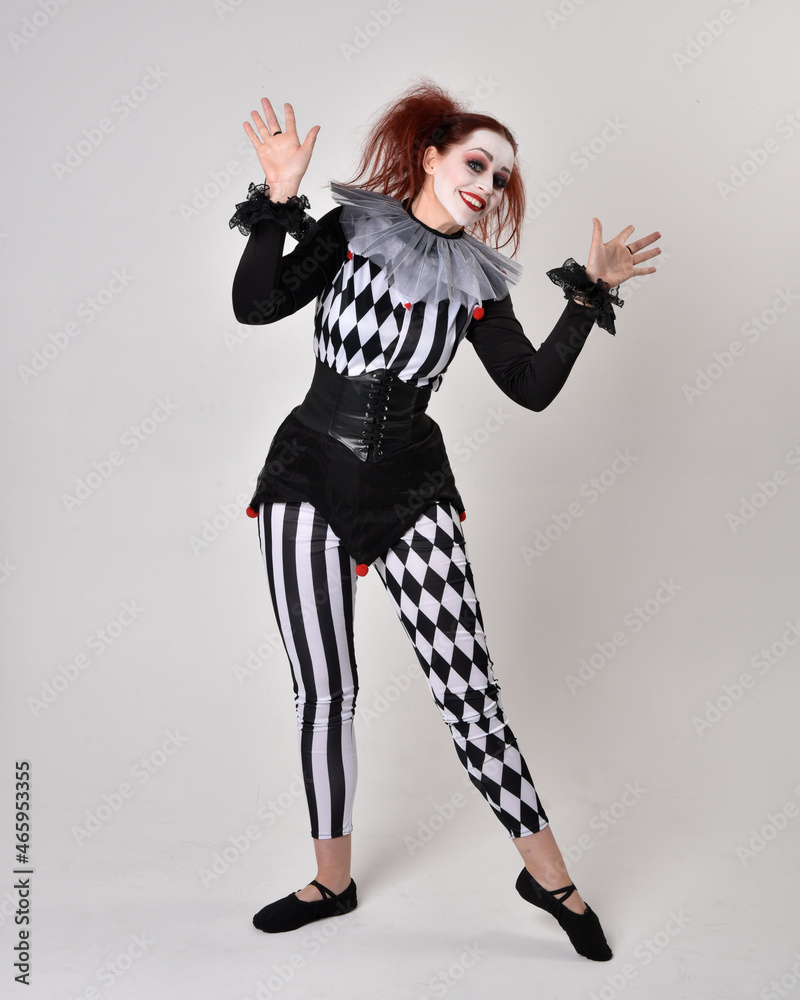 Full length portrait of red haired girl wearing a black and white clown jester costume, theatrical circus character. Standing pose isolated on studio background. Full length portrait of red haired girl wearing a black and white clown jester costume, theatrical circus character. Standing pose isolated on studio background.