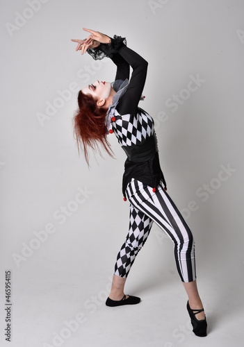 Full length portrait of red haired girl wearing a black and white clown jester costume, theatrical circus character. Standing pose isolated on studio background.