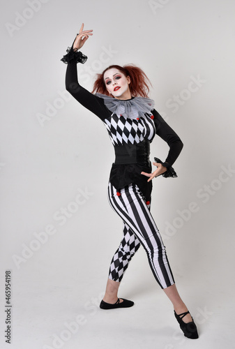 Full length portrait of red haired girl wearing a black and white clown jester costume, theatrical circus character. Standing pose isolated on studio background.