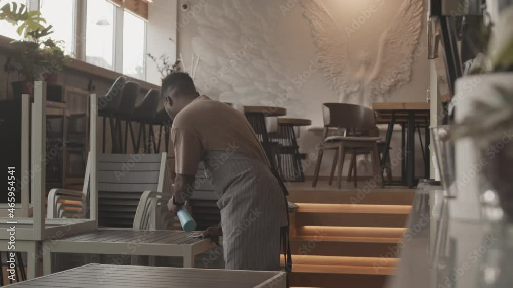 Medium long of young Black man wearing apron spraying cleaner on and wiping table in coffee shop before opening in morning, Biracial female manager coming and talking to him Medium long of young Black man wearing apron spraying cleaner on and wiping table in coffee shop before opening in morning, Biracial female manager coming and talking to him