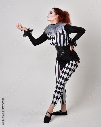 Full length portrait of red haired girl wearing a black and white clown jester costume, theatrical circus character. Standing pose isolated on studio background.