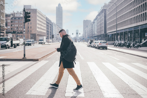 Young bearded caucasian man walking outdoor city crosswalk using smartphone wearing backpack, traveller checking map or commuter going work reading emails photo