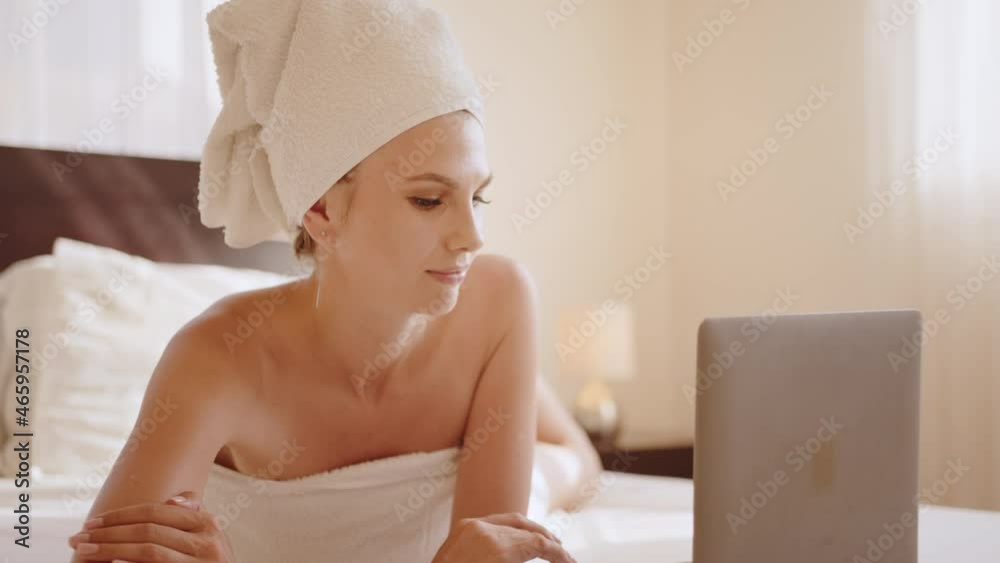 Charming caucasian woman in bath towel using portable laptop during leisure time. Pretty lady lying on comfy bed at hotel room and looking at computer screen. Charming caucasian woman in bath towel using portable laptop during leisure time. Pretty lady lying on comfy bed at hotel room and looking at computer screen.