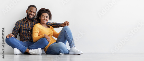 Smiling calm millennial african american couple, owners new apartment sit on floor on gray wall background