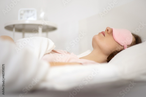 Young beautiful woman sleeping in bed with eye mask in the morning. Woman resting in comfortable white bed, lying on soft pillow, orthopedic mattress, enjoying good sleep.