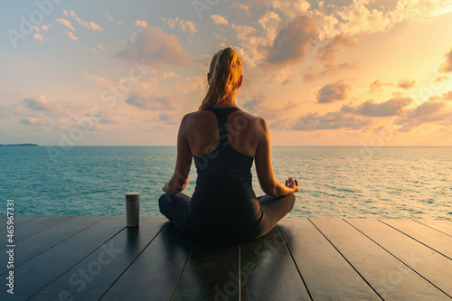 Young woman meditating in a black clothes for yoga  on a wooden deck on the top by the sea. There is a thermo mug nearby.On the background  colorful sunrise sky and sea view.
