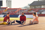 A group of young athletes is sitting on the race track and chatting while warming-up before a training at the stadium. Sport, athletics, athletes