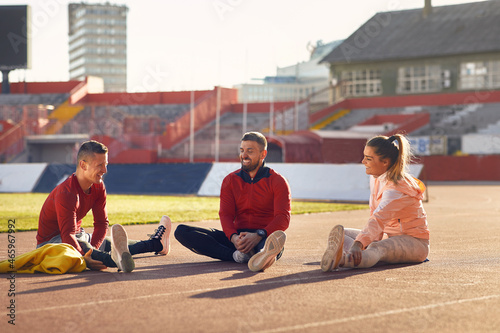A group of young athletes is sitting on the race track and chatting while warming-up before a training at the stadium. Sport, athletics, athletes