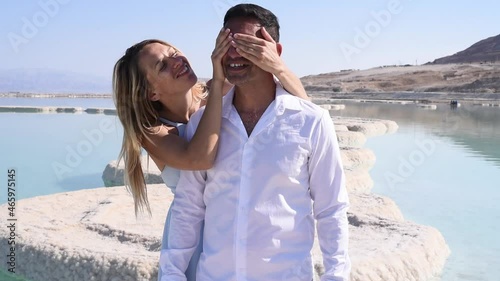 Romantic couple on white beach of Dead sea.  Woman covers man's eyes . Face full of happiness and smile. Marriage on blue sea on paradise. Love story in summer.