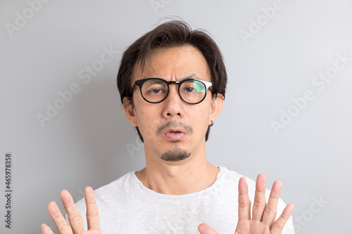 Adult Asian man with eyeglasses raising hand with disagreement on grey background.