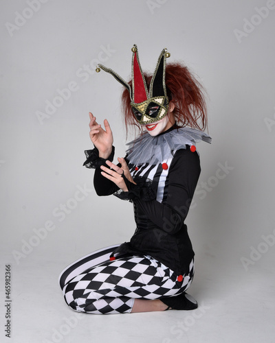 Full length portrait of red haired girl wearing a black and white clown jester costume, theatrical circus character. Sitting down on floor, isolated on studio background.