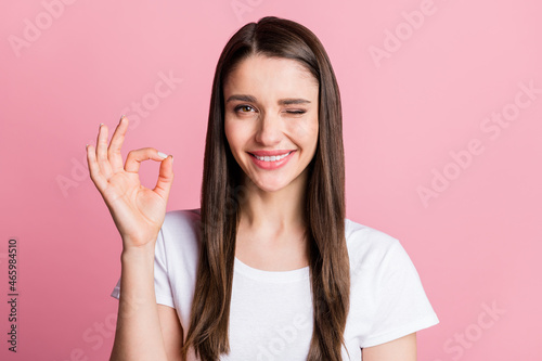 Portrait of attractive cheerful girl showing ok-sign advert winking isolated over pink pastel color background