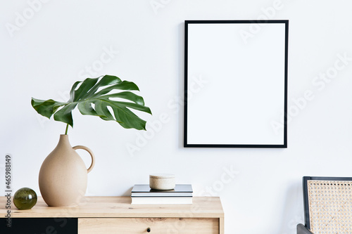 Stylish interior of living room with mock up poster frame, wooden commode, book, tropical leaf in ceramic vase and elegant personal accessories. Minimalist concept of home decor. Template.