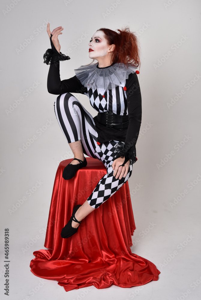 Full length portrait of red haired girl wearing a black and white clown jester costume, theatrical circus character. Sitting down on chair, isolated on studio background. Full length portrait of red haired girl wearing a black and white clown jester costume, theatrical circus character. Sitting down on chair, isolated on studio background.