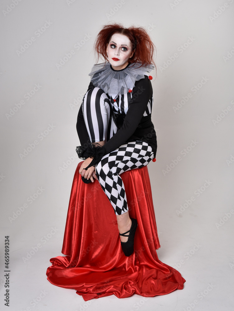 Full length  portrait of red haired  girl wearing a black and white clown jester costume, theatrical circus character.  Sitting down on chair, isolated on  studio background.