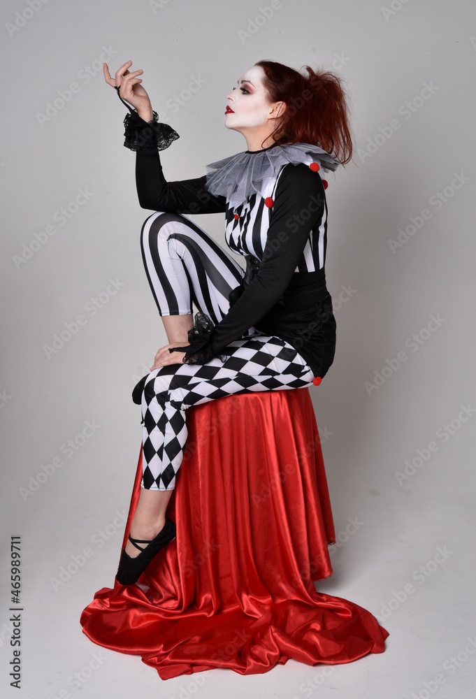 Full length portrait of red haired girl wearing a black and white clown  jester costume, theatrical circus character. Sitting down on chair,  isolated on studio background. Stock Photo