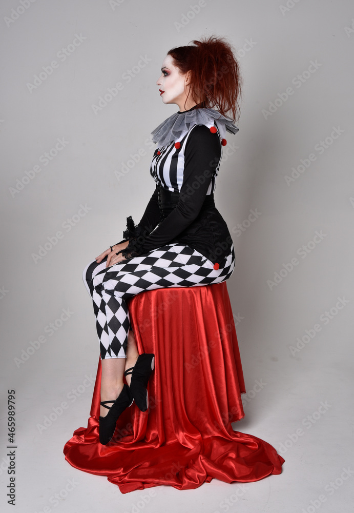 Full length portrait of red haired girl wearing a black and white clown jester costume, theatrical circus character. Sitting down on chair, isolated on studio background. Full length portrait of red haired girl wearing a black and white clown jester costume, theatrical circus character. Sitting down on chair, isolated on studio background.
