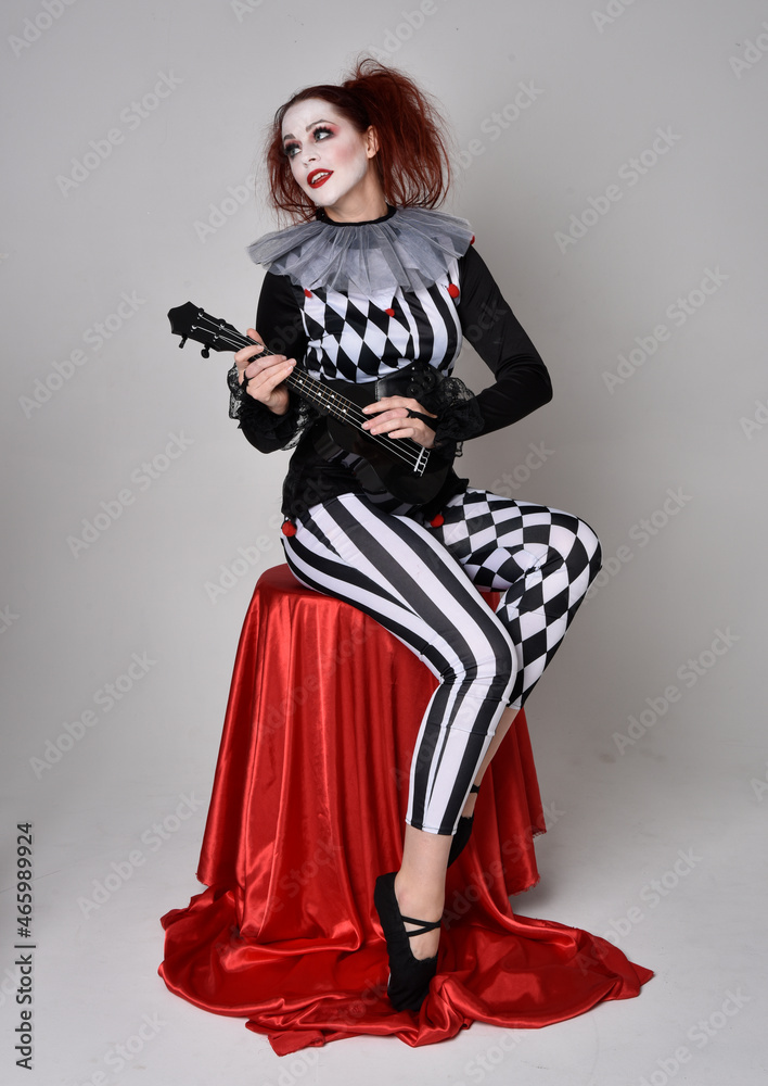 Full length portrait of red haired girl wearing a black and white clown jester costume, theatrical circus character. Sitting down on chair, isolated on studio background. Full length portrait of red haired girl wearing a black and white clown jester costume, theatrical circus character. Sitting down on chair, isolated on studio background.