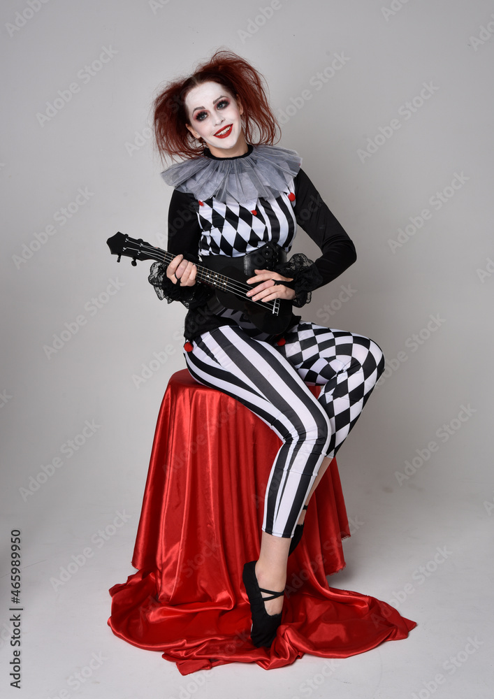 Full length portrait of red haired girl wearing a black and white clown jester costume, theatrical circus character. Sitting down on chair, isolated on studio background. Full length portrait of red haired girl wearing a black and white clown jester costume, theatrical circus character. Sitting down on chair, isolated on studio background.