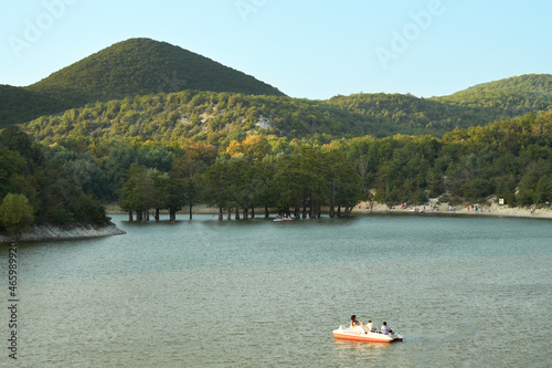 Catamaran boat trip on the lake with swamp cypresses growing out of the water. Numerous tourists walk along the shore. Copy space.