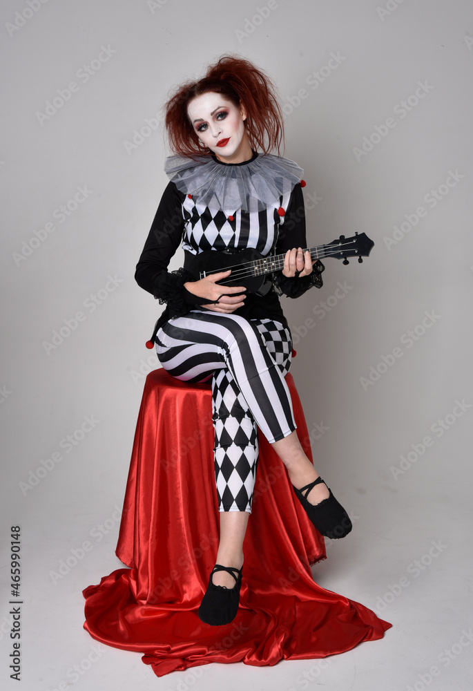 Obraz premium Full length portrait of red haired girl wearing a black and white clown jester costume, theatrical circus character. Sitting down on chair, isolated on studio background.