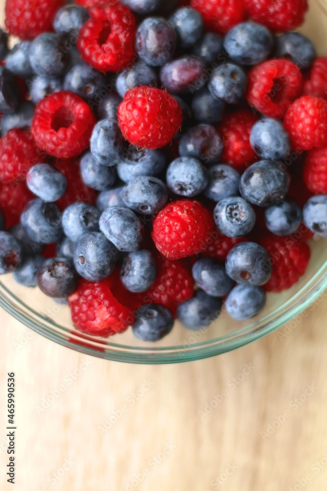 Glass bowl filled with raspberries and blueberries on wooden table. Top view.