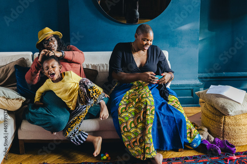 Mother braiding her child's hair while her other mother texts on a smartphone © Lisa Weatherbee