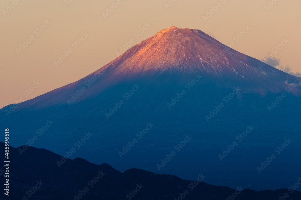Evening light from the setting sun shining on the peak of Mt. Fuji as the sun slowly dips below the horizon as seen from shoreline of Shizuoka Prefecture, Japan.