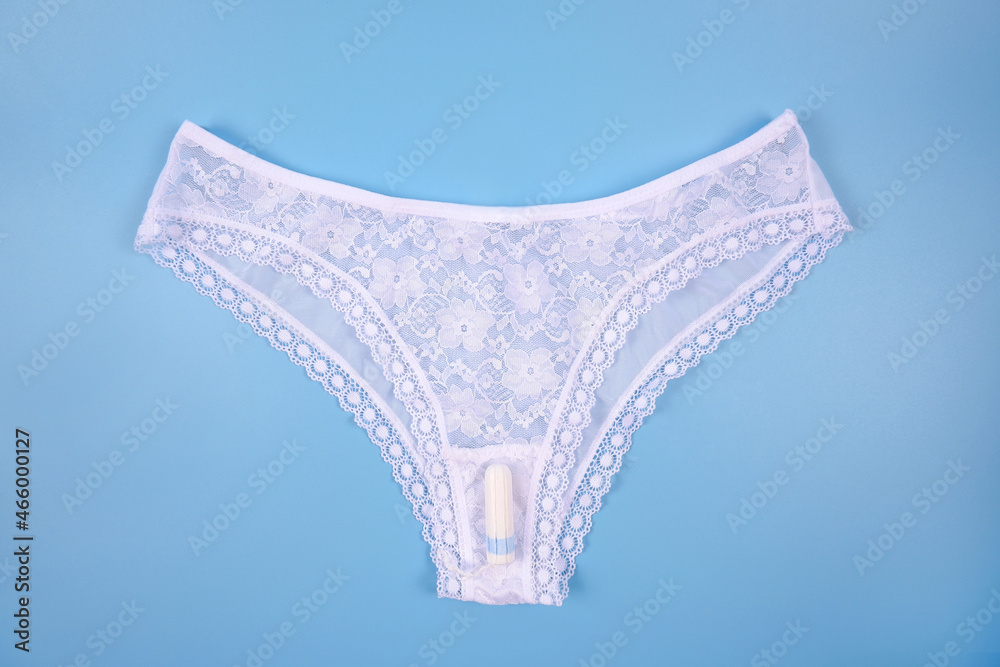 On white sexy lace panties there is a hygienic tampon for women on a blue background. The concept of clothing on critical days, cleanliness during menstruation, feminine hygiene and health.Copyspace - obrazy, fototapety, plakaty 