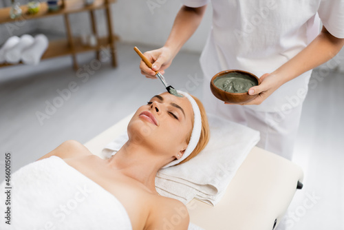 beautician applying clay mask on face of woman lying on massage table