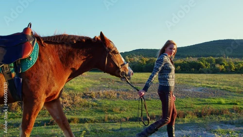 European long-haired girl leads chestnut horse by reins along hill. Panorama, landscape with mountain, wooded steppe. Young woman going with palfrey in countryside. Horsewoman walks bay stallion
