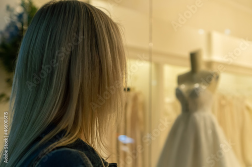 Close up young blonde woman stands at the window of a wedding shop and looks at wedding dresses. Concept of a young woman who wants and dreams of buying a bride's wedding dress