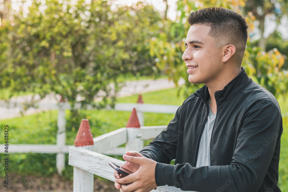 enterprising young man next to the white wooden fence with his cell phone in his hands looking up, happy thinking about his achievements, with a beautiful sunset in the background. enterprising young man next to the white wooden fence with his cell phone in his hands looking up, happy thinking about his achievements, with a beautiful sunset in the background.