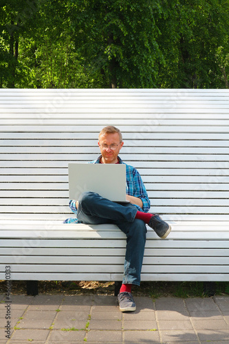 A man with a white laptop on a park bench. The man with glasses and a shirt is a positive freelancer. Vertical photo.