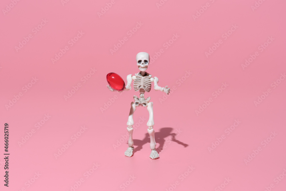 a human skeleton holds a red headache pill in its hands. A trending image on a pink background for the Halloween celebration. Conceptual image about the dangers of medicines. a human skeleton holds a red headache pill in its hands. A trending image on a pink background for the Halloween celebration. Conceptual image about the dangers of medicines.