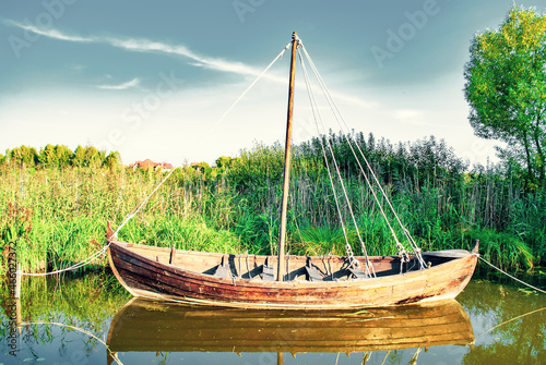 Old wooden boat on lake, side view. Wood rowboat moored near shore by blue dramatic sky