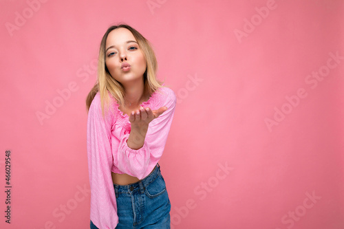 Photo of young positive happy beautiful blonde woman with sincere emotions wearing pink blouse isolated on pink background with copy space and giving kiss