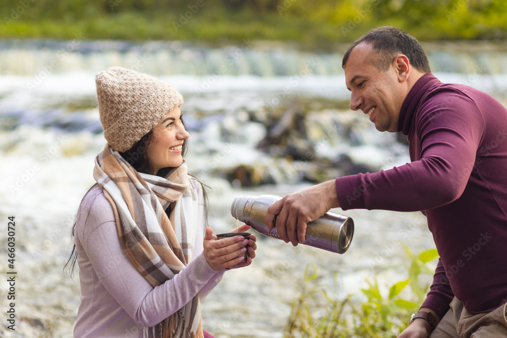 A beautiful young couple is resting on the banks of an autumn river and warm each other up with a hot drink from a thermos. A beautiful young couple is resting on the banks of an autumn river and warm each other up with a hot drink from a thermos.