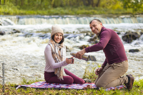 A beautiful young couple is resting on the banks of an autumn river and warm each other up with a hot drink from a thermos. © BY-_-BY