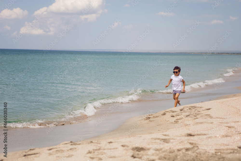 boy child in striped shorts and a white T-shirt walks on sandy beach and in sunglasses