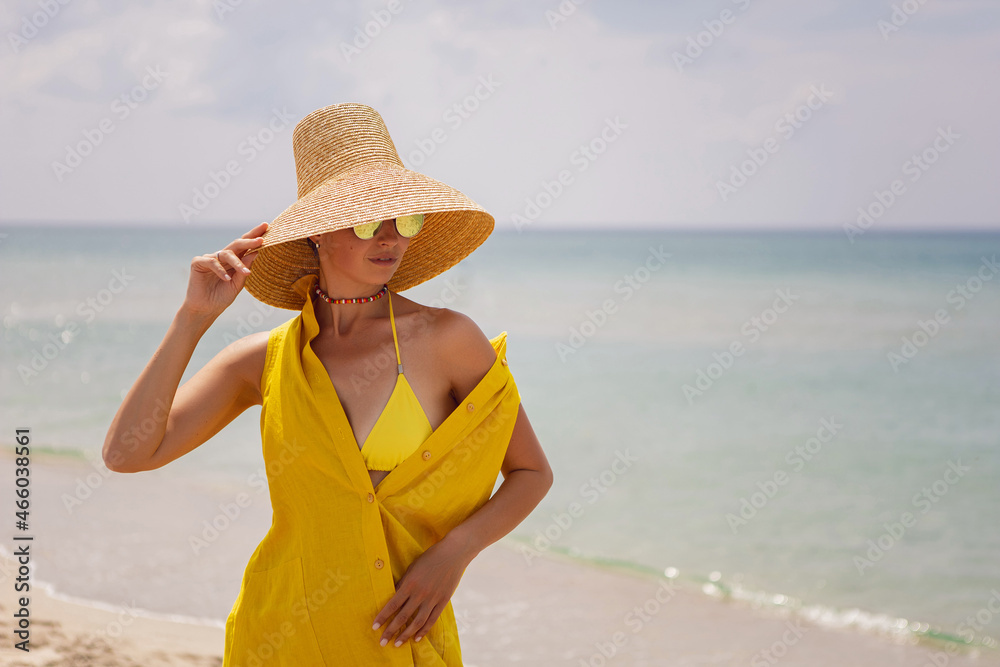 young fashionable woman in a yellow hat dress and sun glasses is posing on beach in summer on vacation