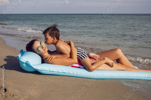 mom and son are lying on the beach on inflatable blue mattress during the summer vacation