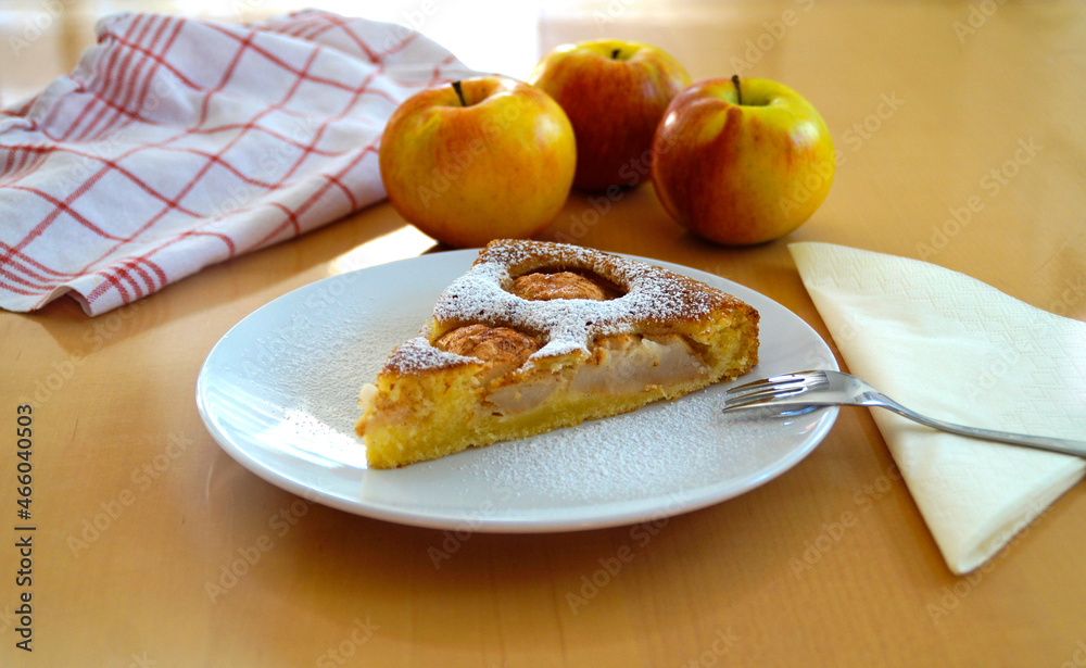 a piece of delicious home-made apple pie (Apfelkuchen) sprinkled with powdered sugar on a plate with three large apples and a kitchen towel in the background a piece of delicious home-made apple pie (Apfelkuchen) sprinkled with powdered sugar on a plate with three large apples and a kitchen towel in the background